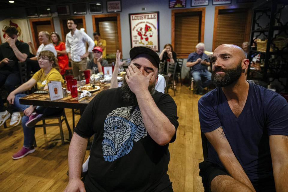 Group of people inside a restaurant, two men in the foreground appear emotional with one covering his face and the other touching his chin