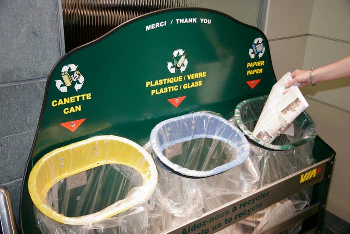 A person's hand is placing paper into a recycling bin with separate compartments for cans, plastic/glass, and paper