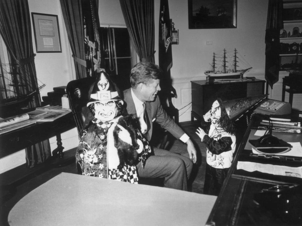 President John F. Kennedy sits behind his desk, laughing as his children Caroline and John Jr. show him their Halloween costumes in the Oval Office of the White House.