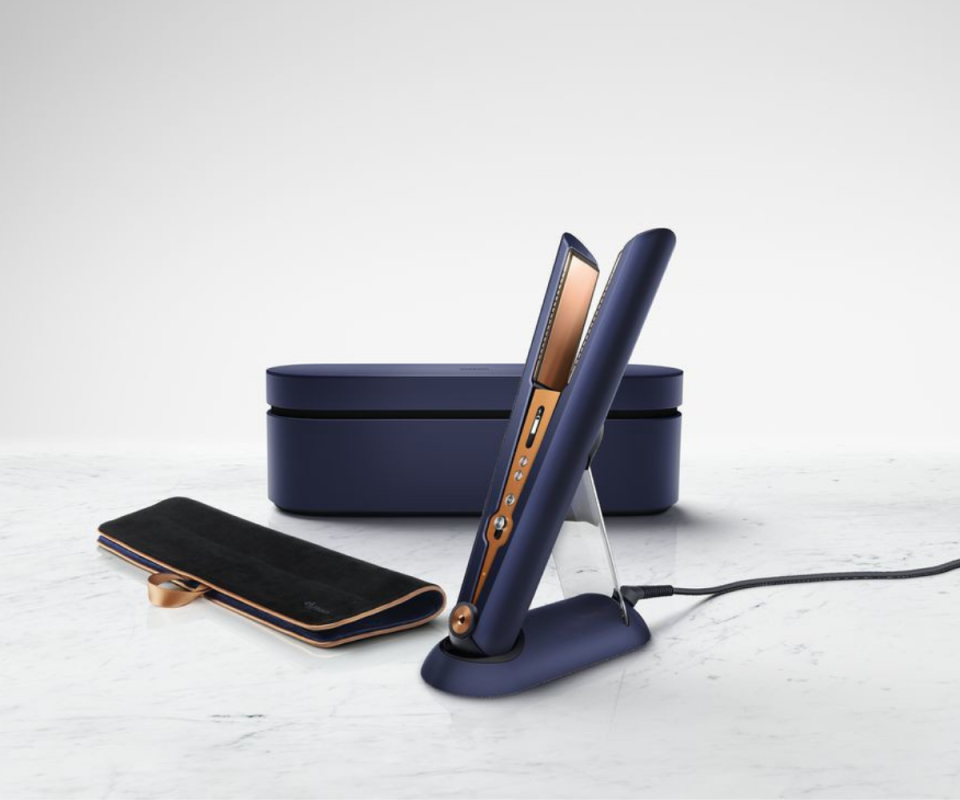 Dyson's Prussian blue and orange copper hair straightener on a stand with a black heat mat and blue box behind on a grey marble backdrop.