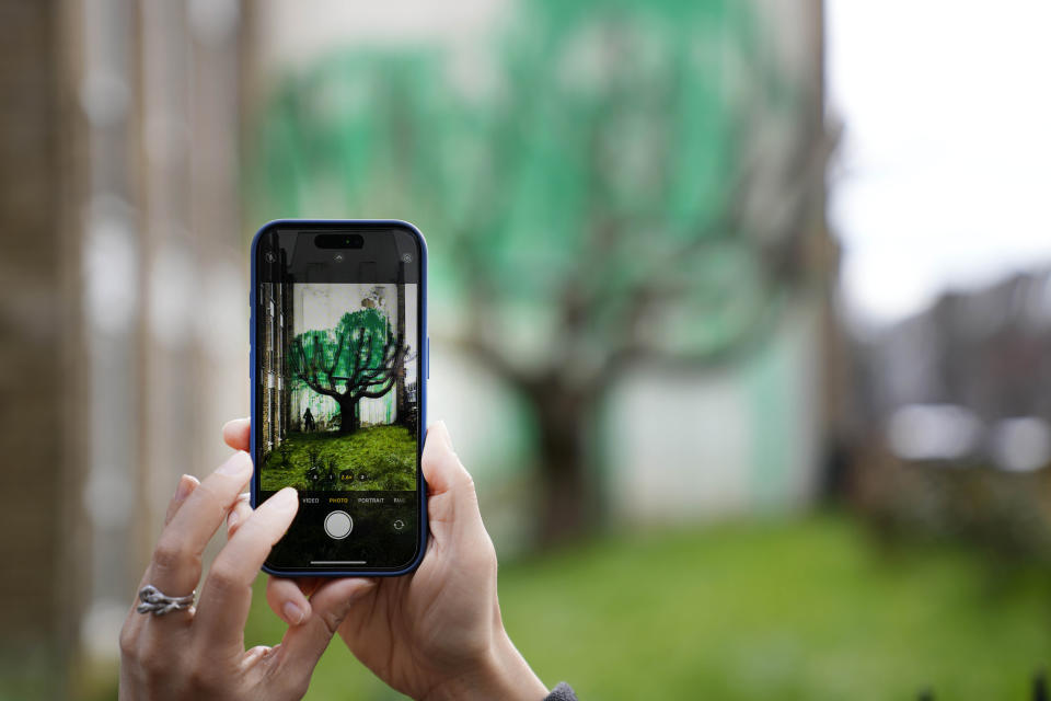 A woman takes a picture on her phone of a new Banksy painting on a wall in London, Monday, March 18, 2024. A new Banksy mural drew crowds to a London street on Monday, even before the elusive graffiti artist confirmed that the work was his. The artwork in the Finsbury Park neighborhood covers the wall of a four-story building and shows a small figure holding a pressure hose beside a real tree. Green paint has been sprayed across the wall, replicating the absent leaves of the tree, which has been severely cropped. Banksy claimed the work by posting before and after photos of the location on his official Instagram account.(AP Photo/Alastair Grant)