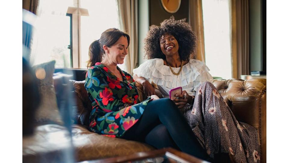 Two beautiful women are sitting comfortably in a luxury hotel, laughing. One of the women is holding a mobile phone in her hand.