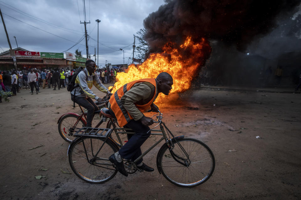 Men ride their bicycles past a roadblock of burning tires in the Kibera neighborhood of Nairobi, Kenya Monday, Aug. 15, 2022. After last-minute chaos that could foreshadow a court challenge, Kenya's electoral commission chairman has declared Deputy President William Ruto the winner of the close presidential election over five-time contender Raila Odinga. (AP Photo/Ben Curtis)