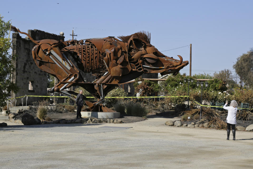 In this Wednesday, Nov. 6, 2019 photo, Paul Witt and his wife Cindy, of Forney, Texas, stop to take pictures beneath a 20-foot sculpture of a boar that still stands in front of the wildfire incinerated Soda Rock Winery in Healdsburg, Calif. If you're worried that wildfires might have created shortages of Northern California's 2019 Cabernet Sauvignon, or even just imparted it with an undesirable smoky flavor, you can relax. The wine is just fine. For now. Despite a late October blaze that raged through one of the world's best-known wine-growing regions. forcing evacuations in two mid-sized towns, wine production in Sonoma County escaped largely unscathed. (AP Photo/Eric Risberg)