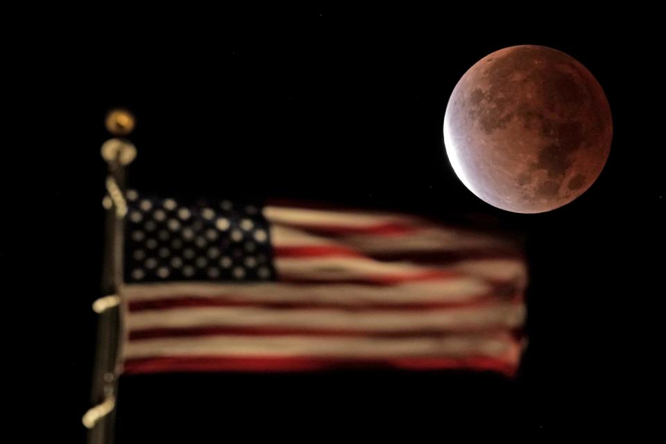 The earth's shadow covers the full moon during a partial lunar eclipse as it sets beyond the U.S. flag on top of a building, Friday, Nov. 19, 2021, in downtown Kansas City, Mo.