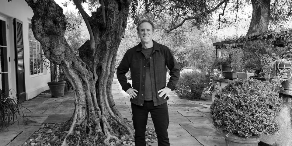 A man stands, hands on hips, by a house and amid greenery.