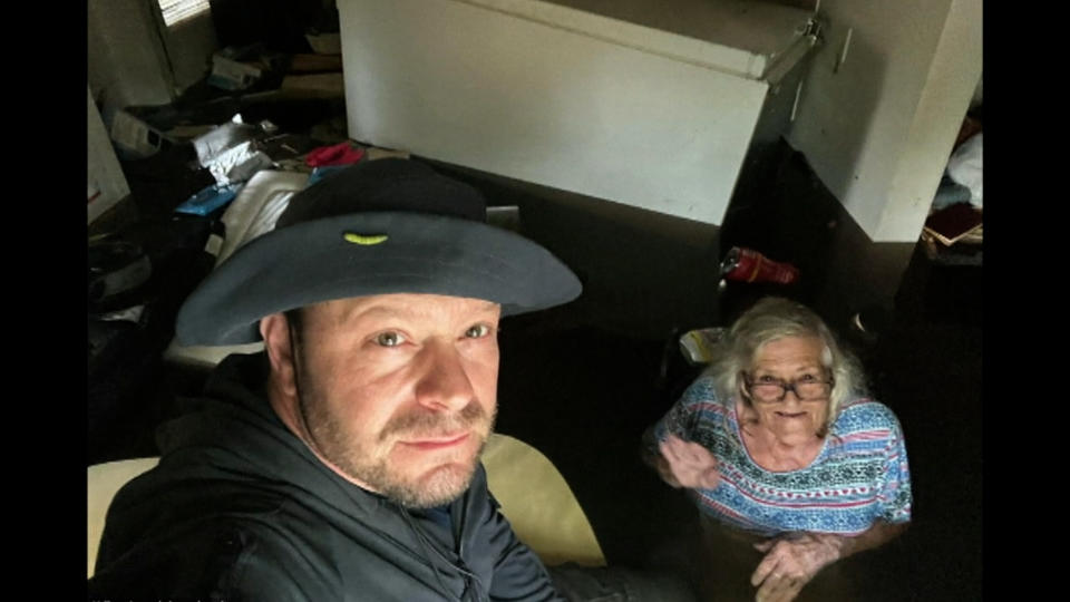 A middle-aged white man in a  gray tilly hat and gray sweater holds a camera aloft for a seflie. His elderly mother with shoulder length white hair smiles, despite being in waist deep brown water inside her home. (JOHNNY LAUDER / Associated Press)