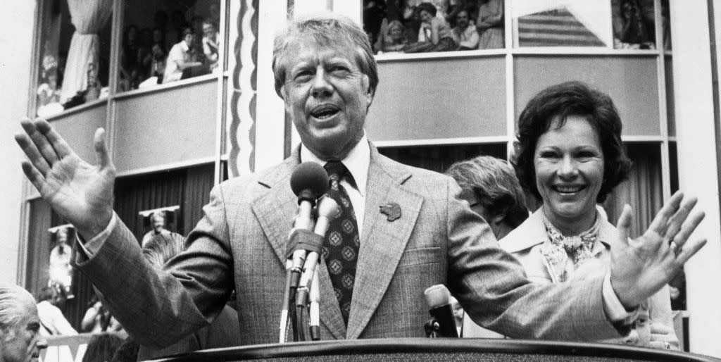 jimmy carter wearing a suit and tie and speaking into a microphone at a podium, standing next to his wife rosalynn, with supporters in stands behind him