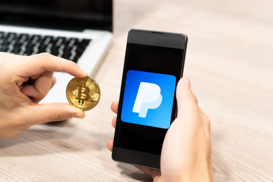 Ein Mann hält eine Bitcoin-Münze neben ein Smartphone mit PaypalLogo auf dem Bildschirm