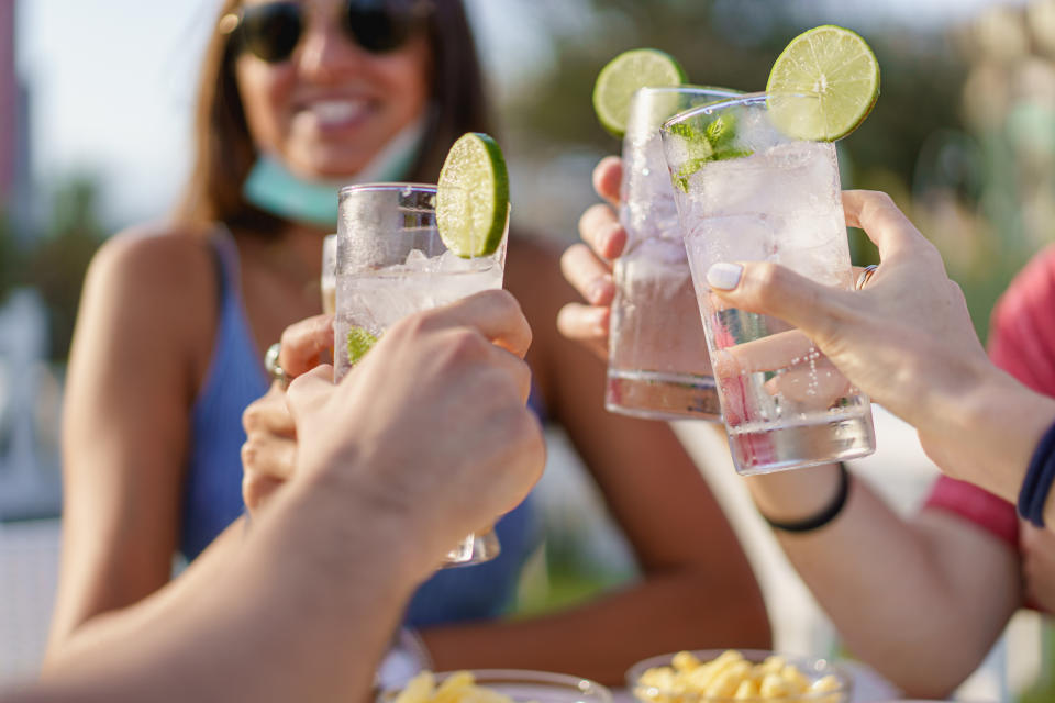 Friends drinking cocktails in an outdoors snack bar restaurant in the summer wearing face mask on to be protected from coronavirus - Happy people cheering with mojito and having fun