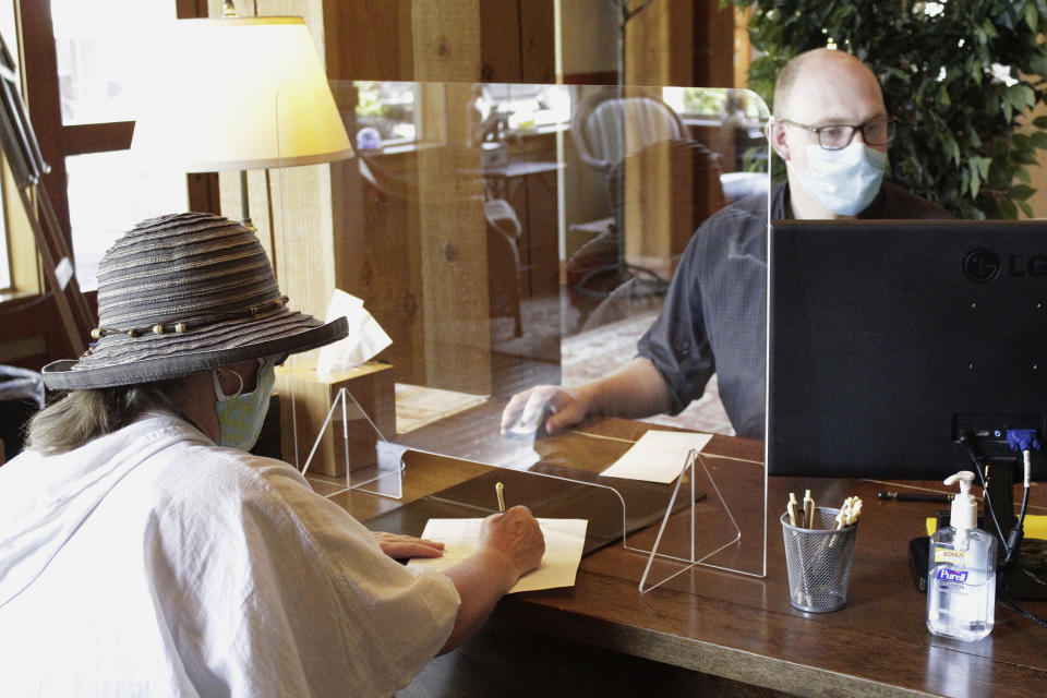 In this Thursday, May 28, 2020, photo, hotel employee Daniel Mathre, right, helps a guest from behind protective plastic at The Ocean Lodge in Cannon Beach, Ore. The Ocean Lodge welcomed guests back starting last week after an extended shut-down of all hotels and vacation rentals in the small beach town to prevent the spread of the new coronavirus. With summer looming, Cannon Beach and thousands of other small, tourist-dependent towns nationwide are struggling to balance fears of contagion with their economic survival in what could be a make-or-break summer. (AP Photo/Gillian Flaccus)