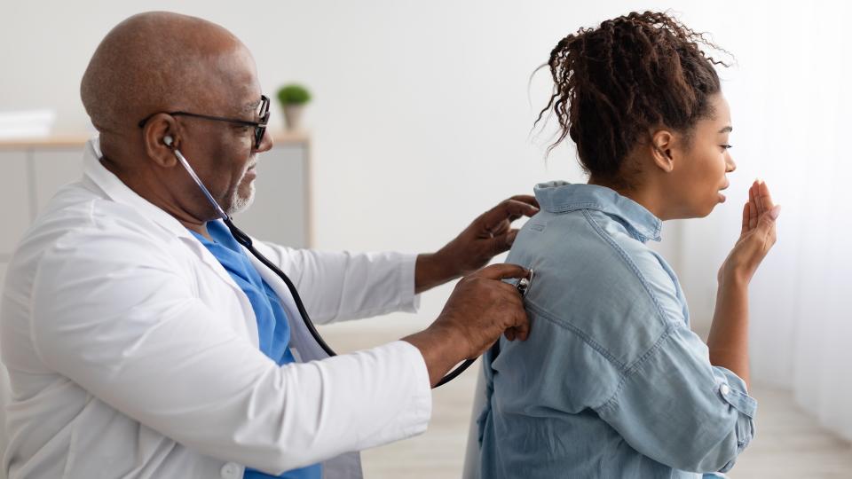 Black male doctor checking breath of female patient using a stethoscope, listening to her lungs from the back. She is holding up a hand to cover her mouth and cough.