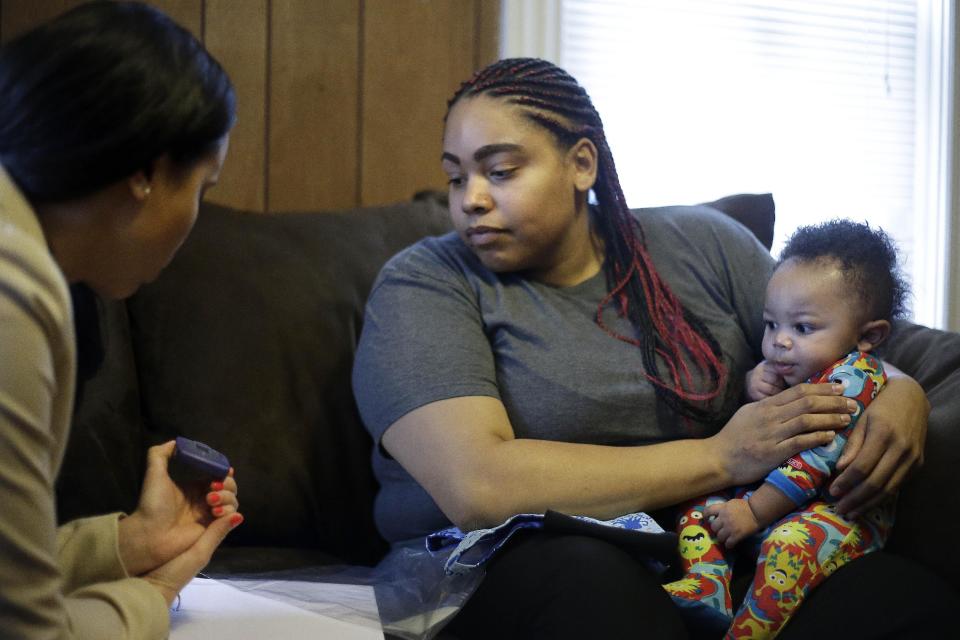 In this Feb. 3, 2014 photo, caseworker and home visitor Stephanie Taveras, left, shows Ashley Cox, center, a digital audio recorder as Cox holds her six-month-old son Jaiven, at the family's home in Providence, R.I. The city has begun an effort to boost language skills for children from low-income families by equipping them with audio recorders that count every word they hear. During home visits, social workers go over the word counts with parents and suggest tips to boost the child’s language skills. (AP Photo/Steven Senne)