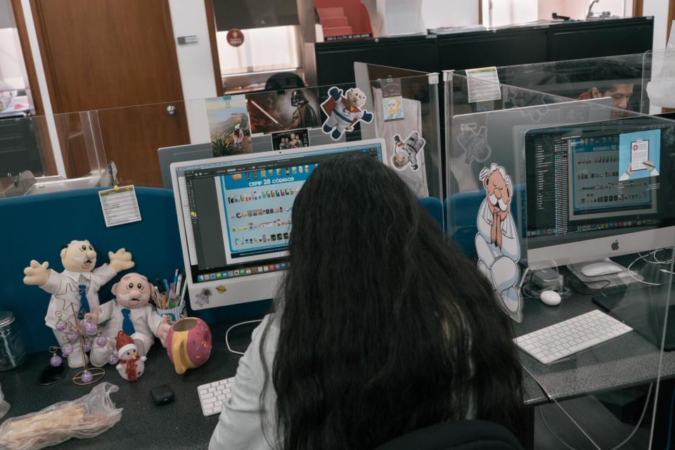 A woman with long dark hair sits at a computer terminal. On the desk are dolls of a man in a lab coat