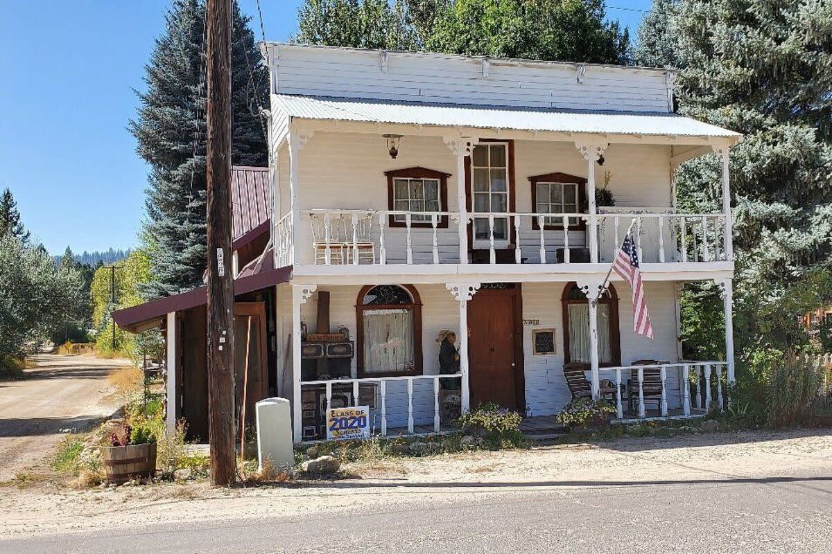 White house in the Boise Basin Museum Historical Town, Idaho City, Idaho, surrounded by trees and plants, paved road in the foreground, dirt road on the left