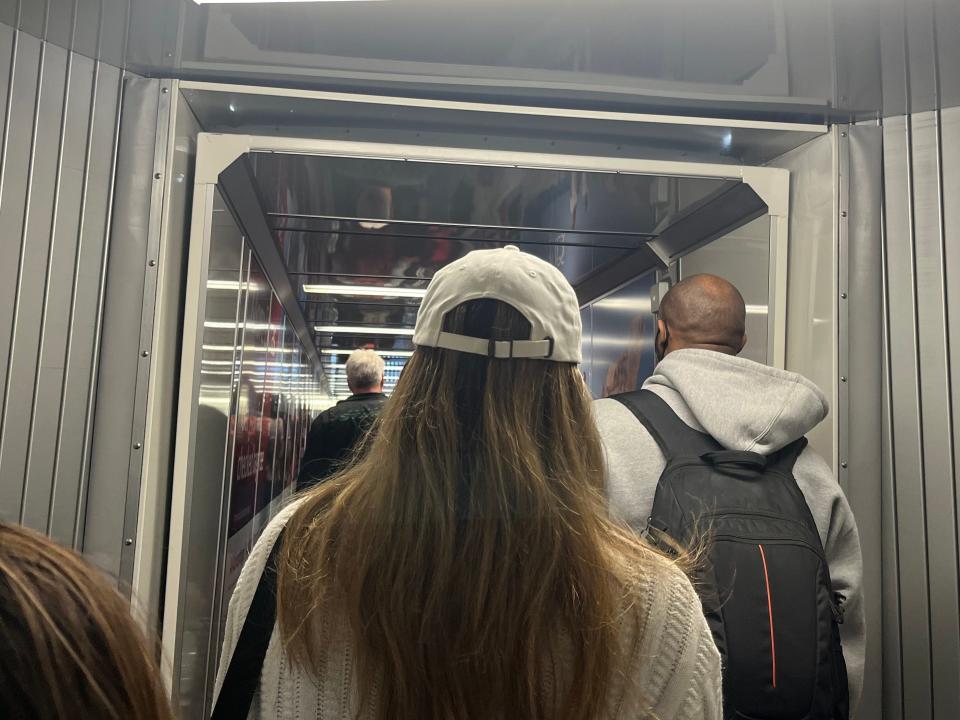 A group of people, including a man in gray sweatshirt and backpack, and a woman in a baseball cap and white sweater, stand on the jetway while waiting to board a flight.