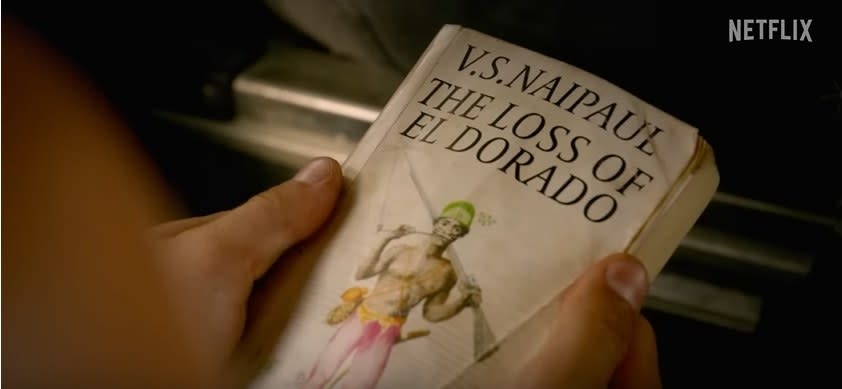 someone out of frame holding a book titled "V S Naipaul the Loss of El Dorado"