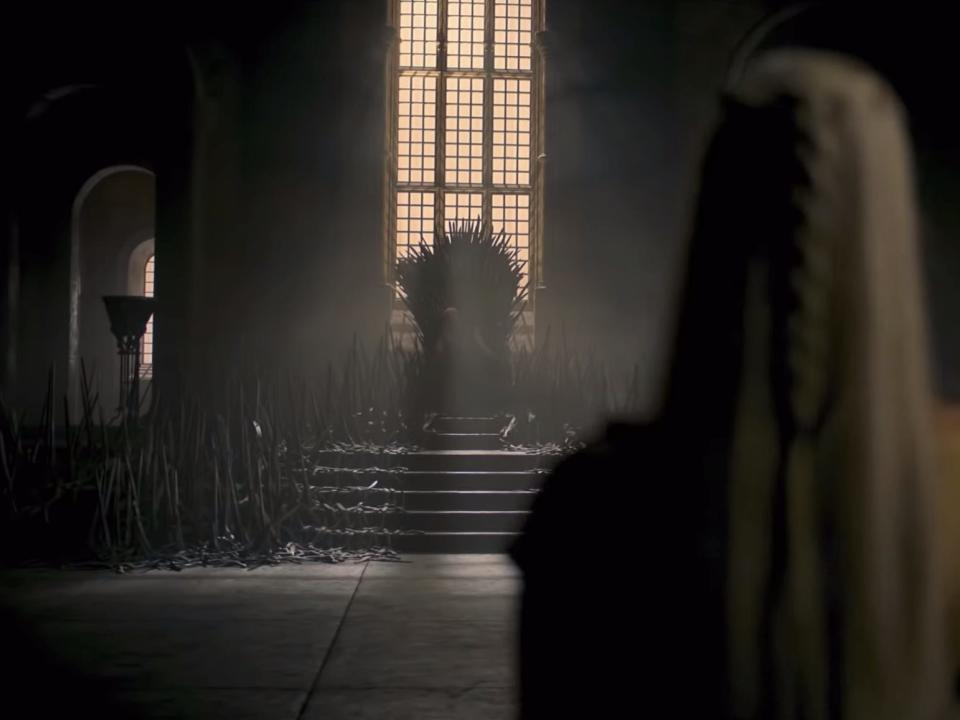 A blonde-haired woman stands in the foreground with the Iron Throne ahead of her.