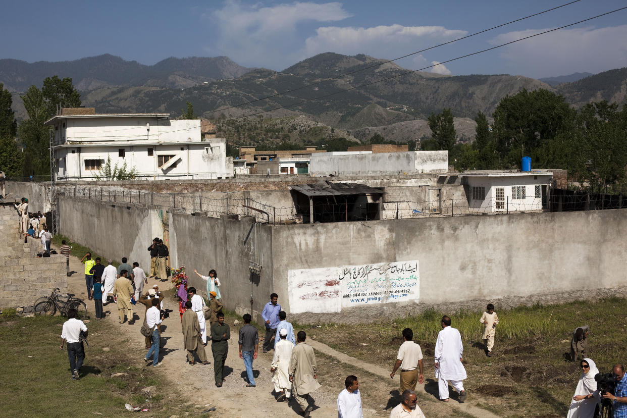 People walk on a dirt path outside Osama Bin Laden's compound, near a concrete wall with barbwire at the top, with mountains visible in the distance.