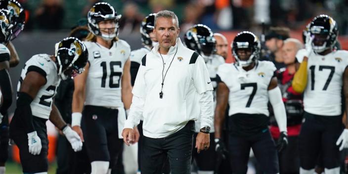 Urban Meyer stands on the field while Jaguars players stand behind him.