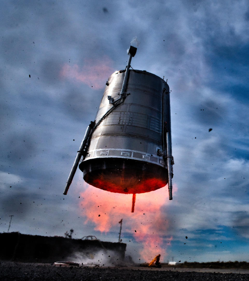 a cylindrical silver rocket stage flies low to the ground, with orange flames erupting from its base.