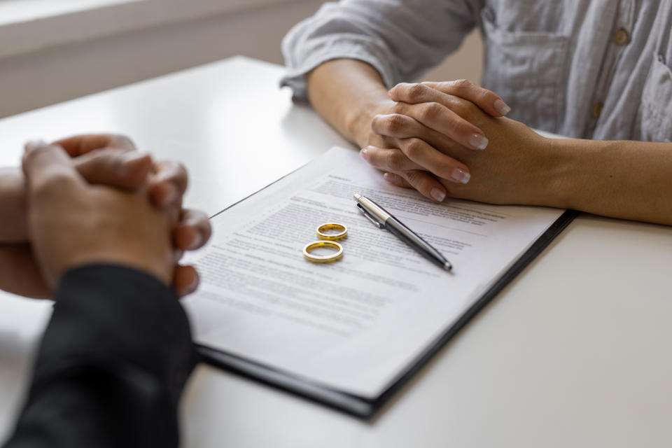 Two individuals are seated at a table with a document, pen, and two wedding rings, suggesting the signing of a marriage or divorce agreement