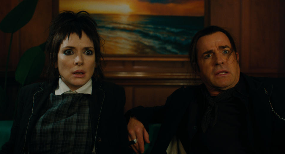 Winona Ryder and Justin Theroux sit on a couch, looking surprised, with a painting of a beach sunset behind them
