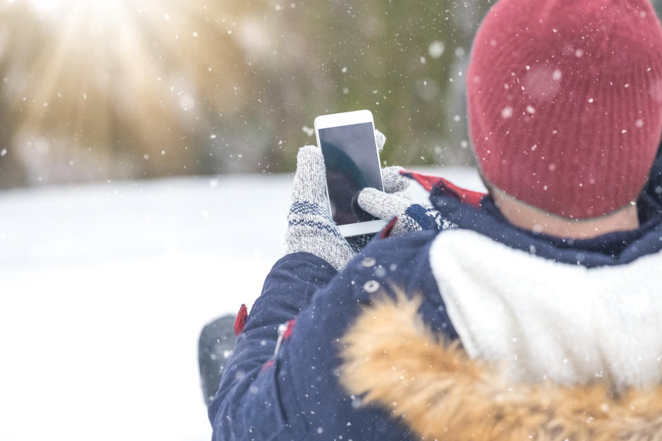 Rear view of man sitting in snow and holding blank screen smart phone.