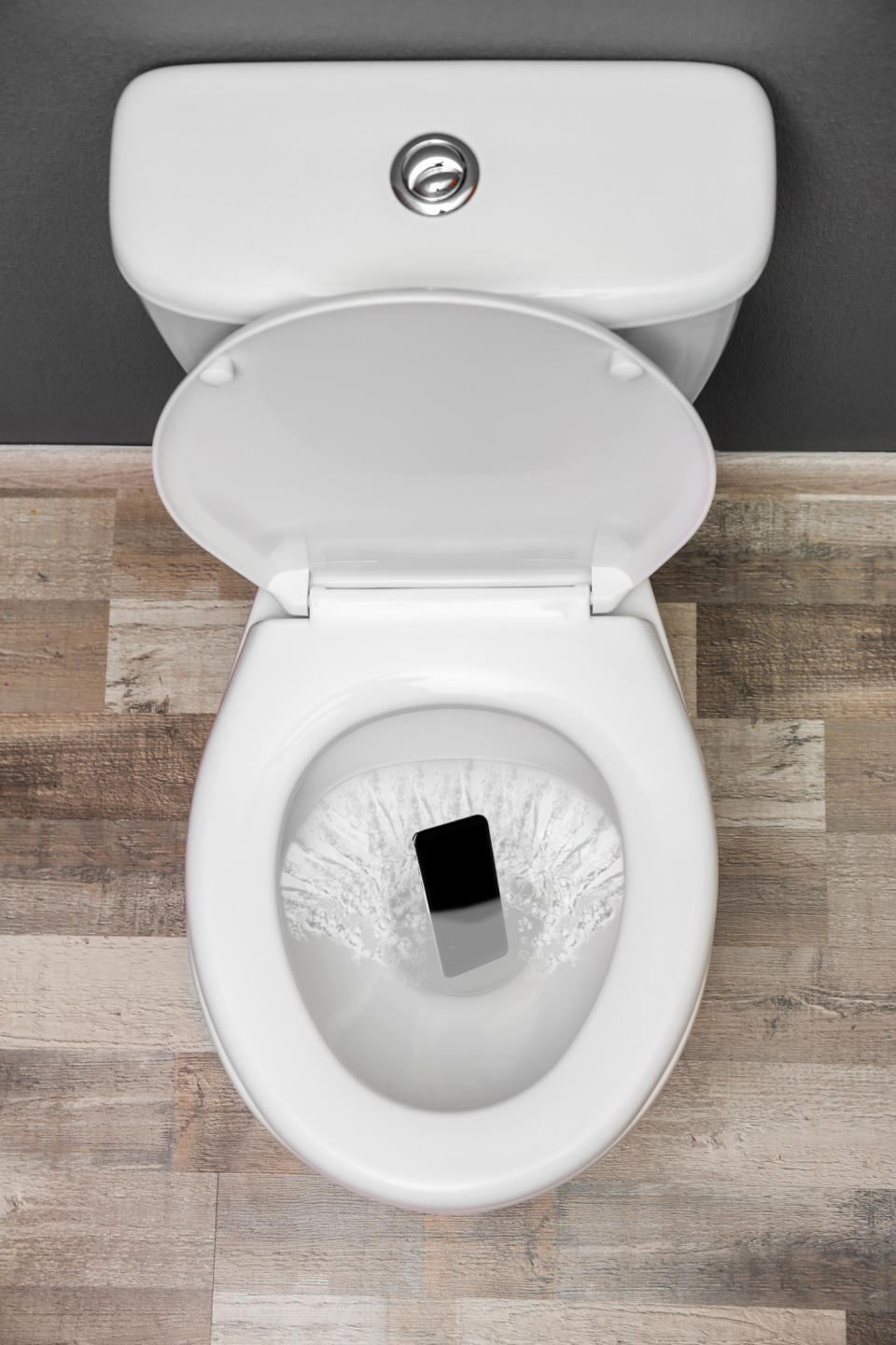 A smartphone is falling into a toilet bowl mid-flush, suggesting an accidental drop