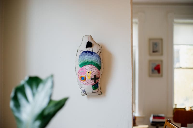 Embroidered pillow hung on wall above stool in light filled apartment.