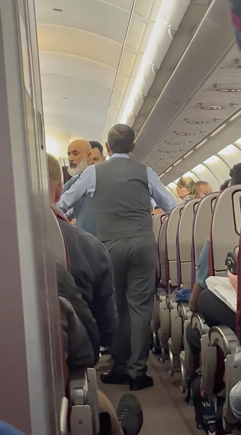 In this image from a video, a passenger, left, speaks with crew members during a flight from Australia to Malaysia, Monday, Aug. 14, 2023. Malaysia Airlines flight MH122 returned on Monday to Sydney, where the passenger was arrested hours later in what police described as an emergency incident. (Velutha Parambath via AP)
