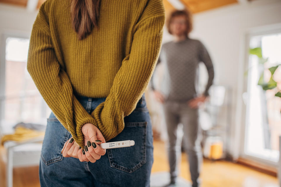 Person with long hair in a sweater and jeans, holding a positive pregnancy test behind their back. Another person in the background, smiling. 