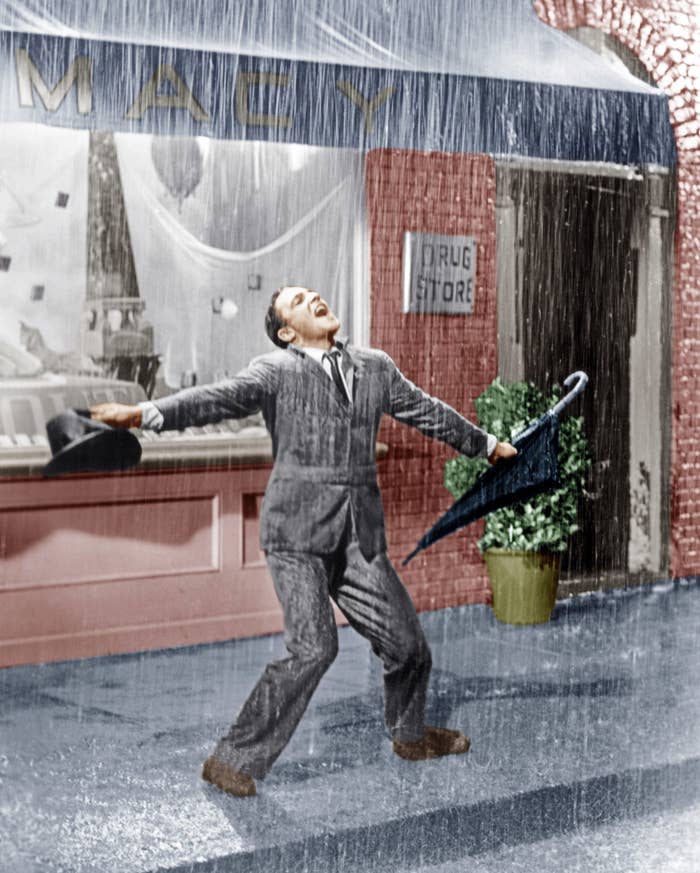 Gene Kelly dances happily in the rain, holding an umbrella and a hat, wearing a suit, in front of a shop window