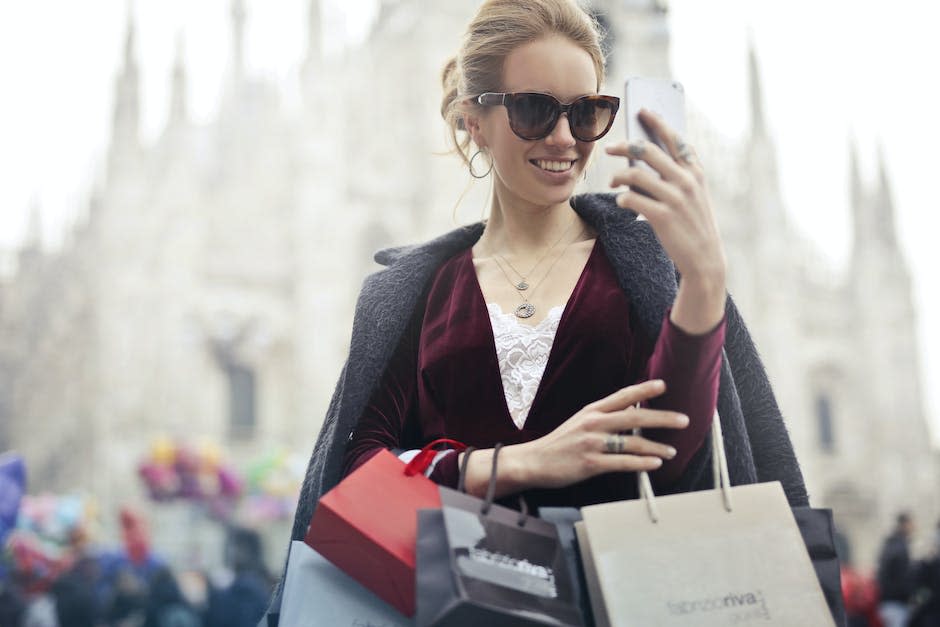 Mujer En Top De Manga Larga Granate Sosteniendo Smartphone Con Bolsas De Compras Durante El Día