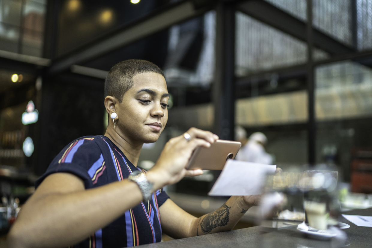 Young woman depositing a check by smartphone while in a cafe, with a blurred background of table, large walls, and windows