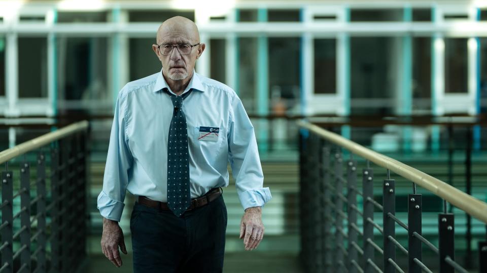 A bald man with glasses and a white goatee is walking across a building walkway. He is smartly dressed, wearing a long-sleeved light shirt, slacks, and a blue tie. He looks worried.