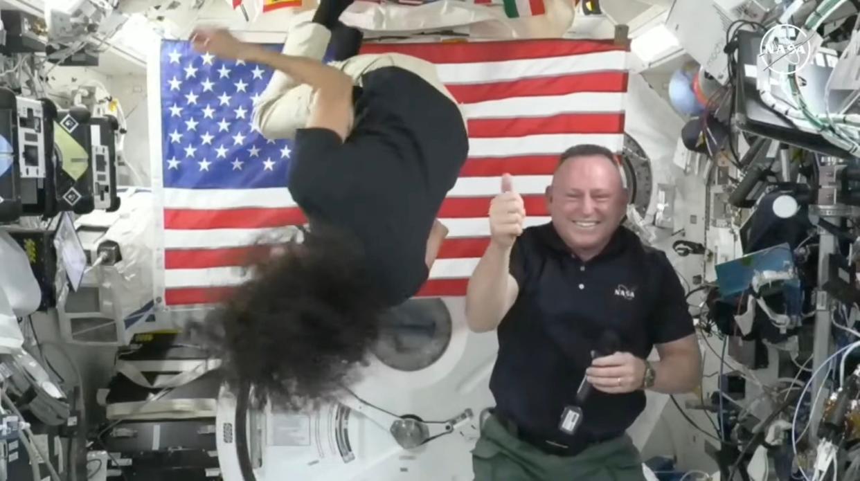 astronaut suni williams upside down flipping near the ceiling of the space station while butch wilmore gives a thumbs up smiling next to her in front of an American flag beside walls covered in gadgets