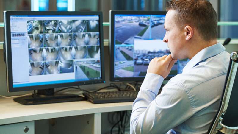 A man in a white Oxford shirt sits, monitoring screens that display footage from surveillance cameras.