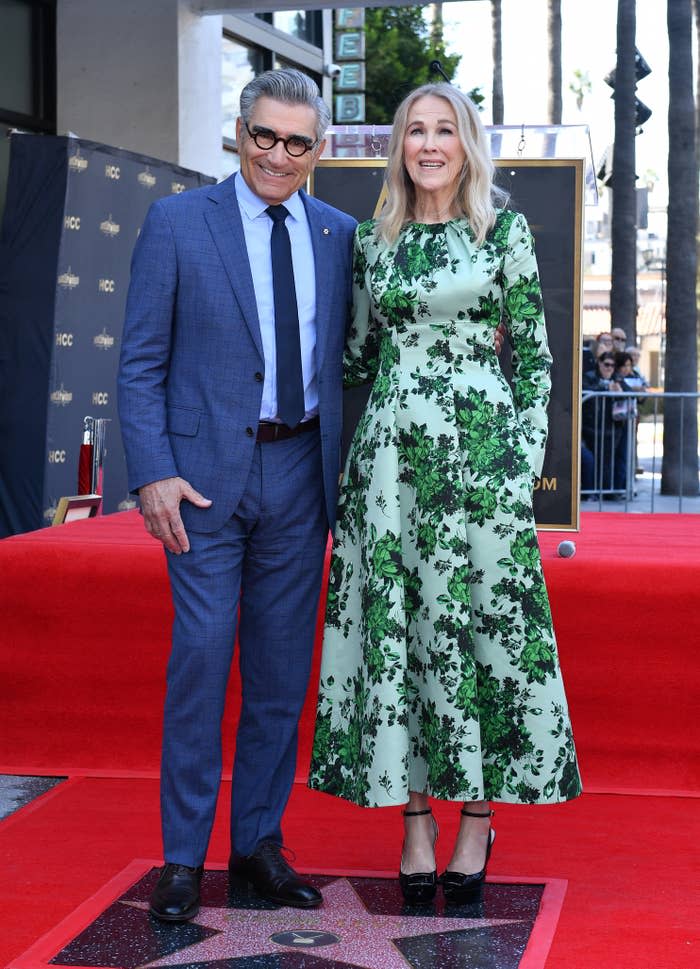 Eugene Levy and Catherine O'Hara stand side by side on a red carpet; she wears a floral dress, he's in a blue suit