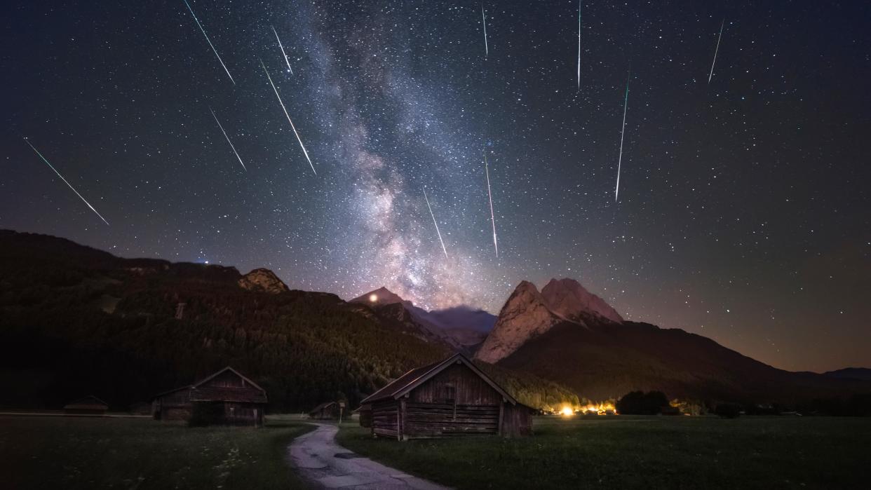  meteors streak through a star filled sky above mountains and small wooden lodges. 