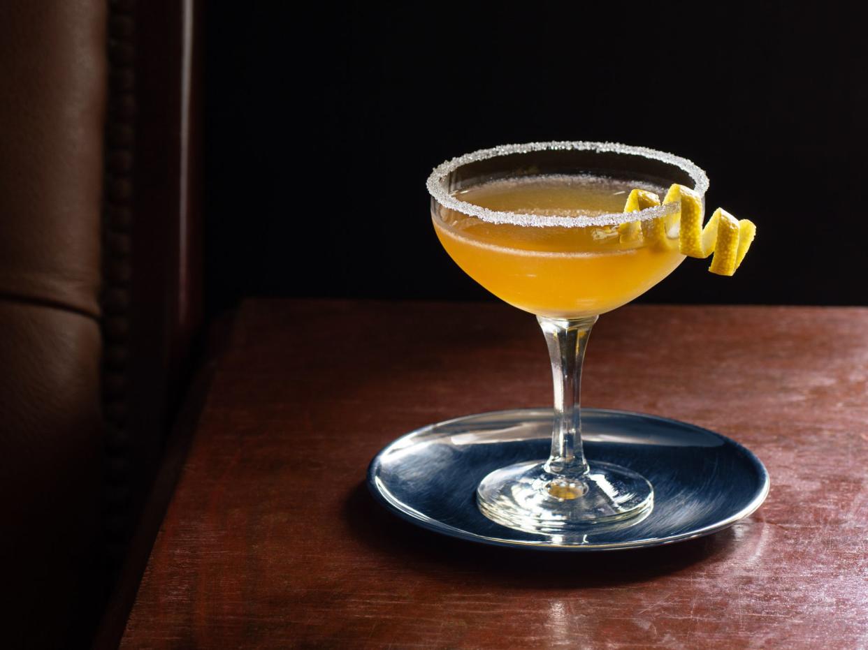 A chilled sidecar cocktail in a coupe glass with a sugared rim on a dark wood table next to a leather seat. This drink is made from cognac, Armagnac, or brandy mixed with an orange liqueur and lemon juice, shaken with ice and strained. Taken in a dark lux