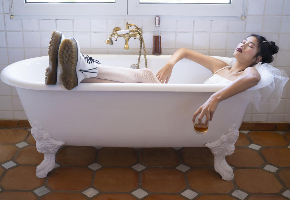 A person wearing white boots and a white dress reclines in a vintage bathtub, holding a glass of brown liquid, appearing relaxed