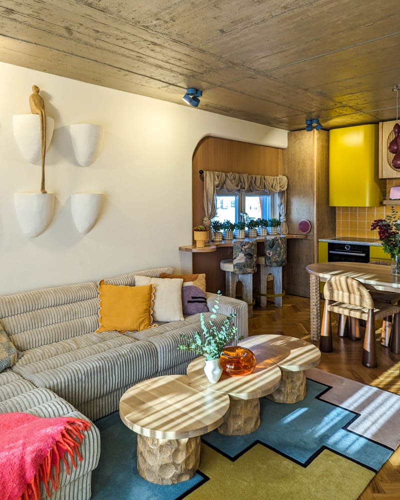 white living room with large sofa, circles coffee table, pops of color, and yellow kitchen in background