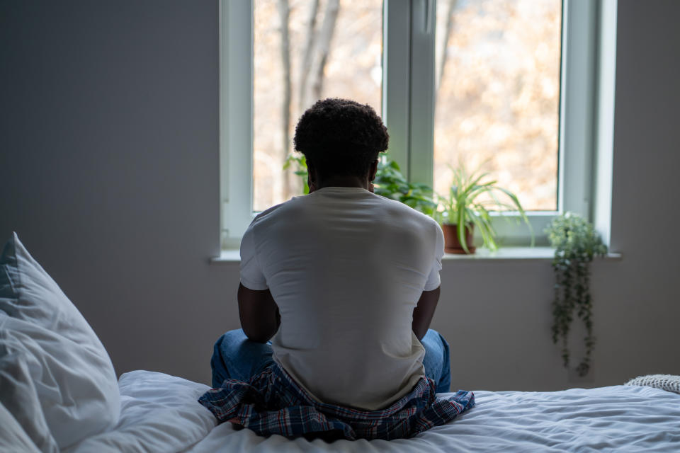 A person sits on the edge of a bed looking out of a window, appearing contemplative. Houseplants adorn the window sill