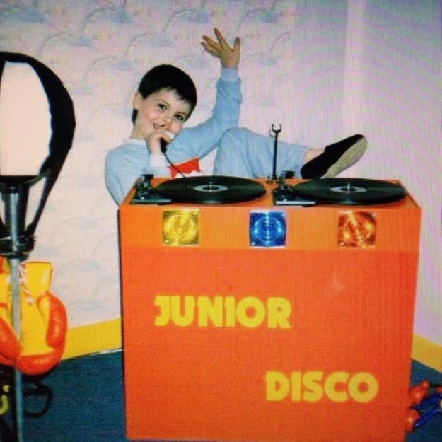 Child sitting at a "Junior Disco" booth with records on top and holding a microphone