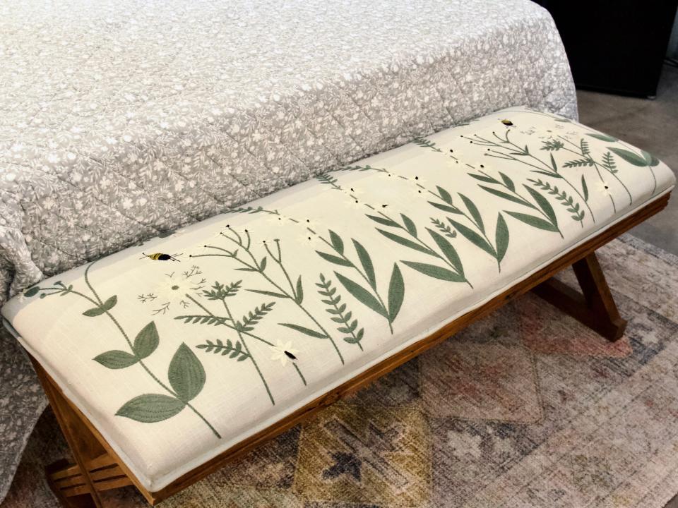 An embroidered wooden bench with flower and bee details at the foot of a bed
