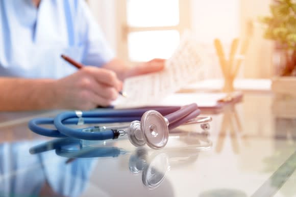 Stethoscope on table with person dressed in scrubs in background.