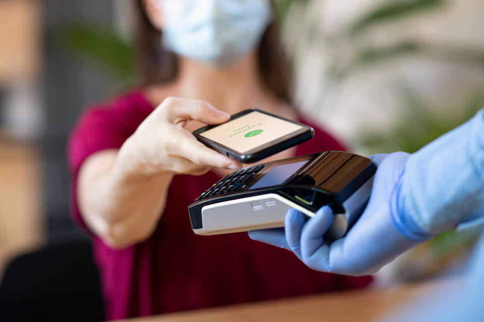 Close up hand of customer paying with smartphone. Cashier hand holding credit card reader machine and wearing protective disposable gloves at bar counter, while client holding phone for NFC payment. Woman wearing face mask while paying bill with mobile phone during Covid-19 pandemic.