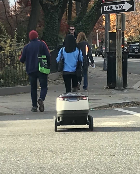 Small white robot on two wheels crossing the street and trailing three people