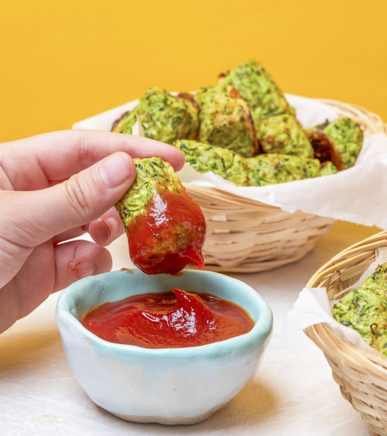 a zucchini tater tot being dipped into a small blue bowl of ketchup in front of a parchment basket of tots